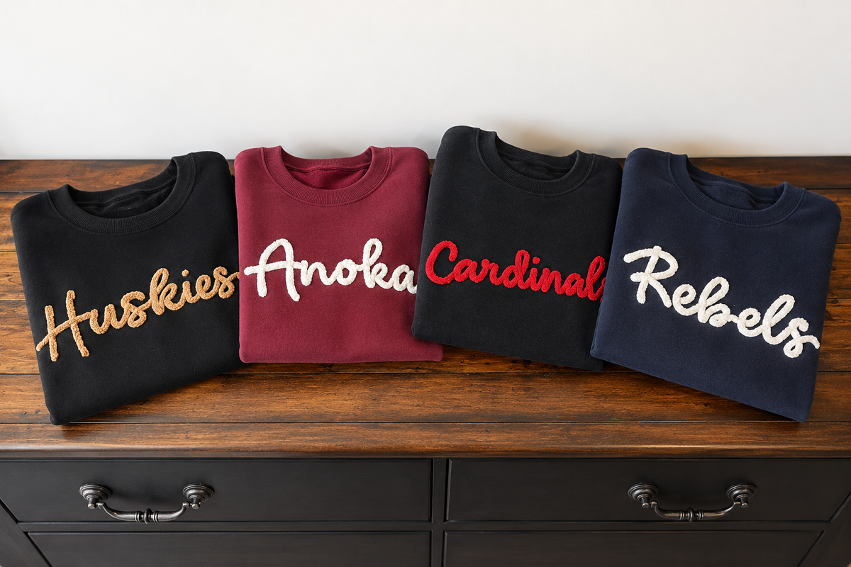 Four folded sweatshirts displayed side by side on a wooden cabinet against a plain white wall. From left to right, the sweatshirts are black with gold 'Huskies' lettering, burgundy with white 'Anoka' lettering, black with red 'Cardinal' lettering, and navy blue with white 'Rebels' lettering.